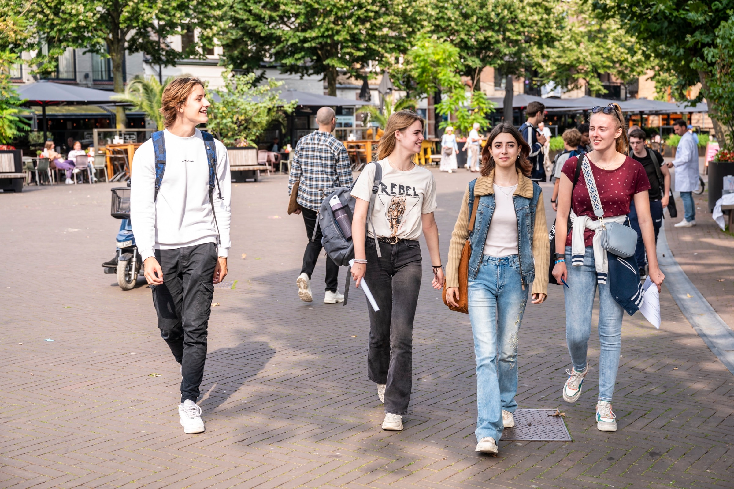 Vier jongeren lopen lachend over een plein in het centrum van Oss, met terrassen en bomen op de achtergrond.