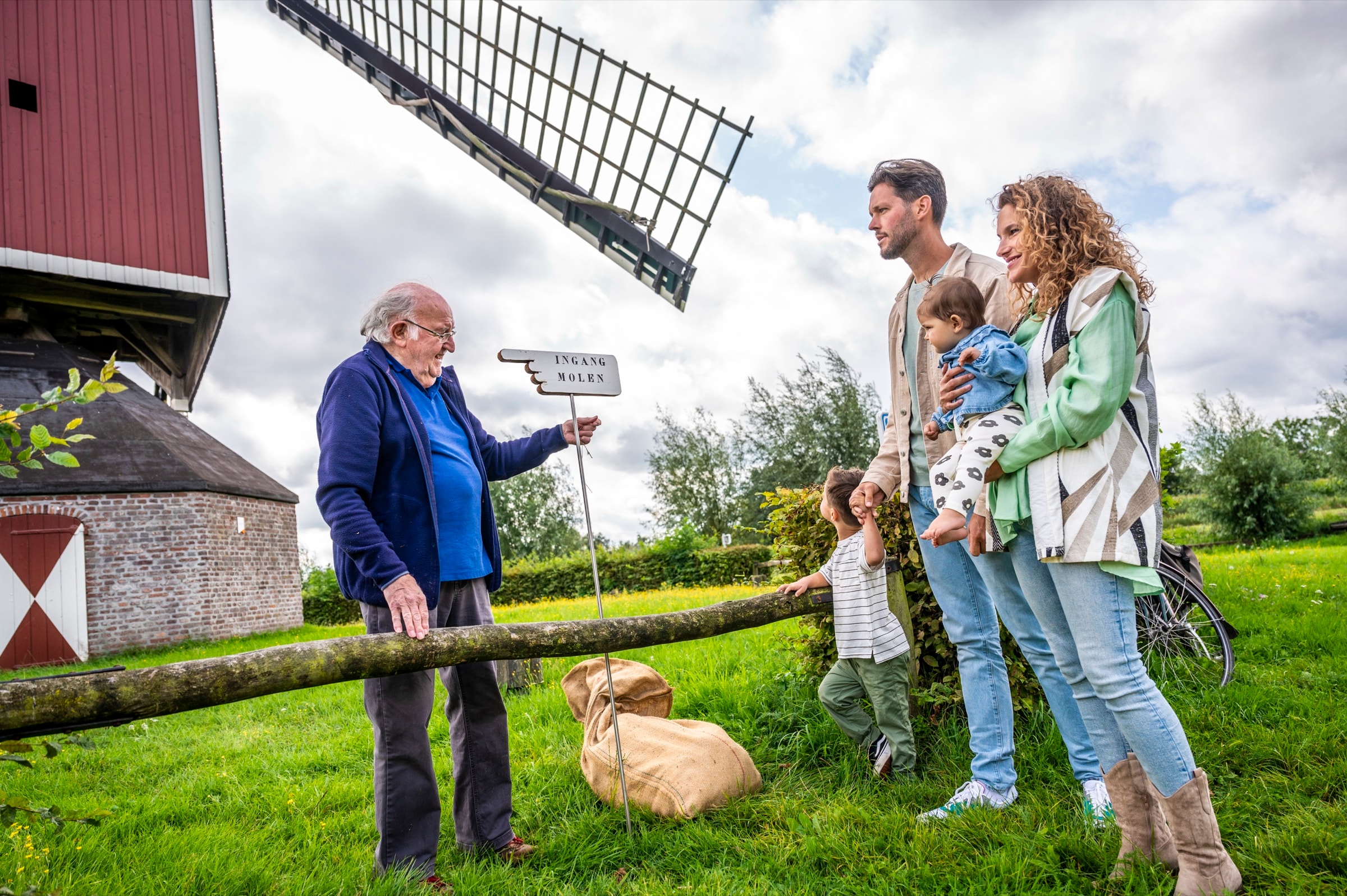 Een man wijst naar een informatiebord bij een molen; een gezin met kinderen kijkt toe.